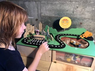 A child with redish golden hair in a black t-shirt looking at a small scale 3D model of the city, with a road, the landfill and heavy vehicles.