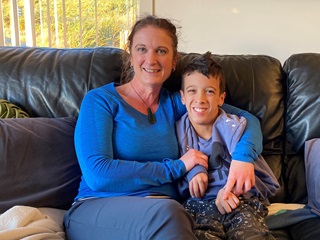 Kris Dahl, dressed in blue, holding her son Cyrus Dahl in an embrace on a couch, both smiling.