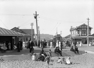 Black and white photo of Lyall Bay foreshore between 1910 and 1914 before the sea wall was constructed, with children playing in the sand, and women in long dresses and men in top hats standing around the tram, tram shelter, and tea rooms.