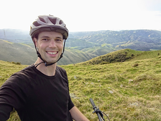 Mark Noyes, taking a selfie with his helmet on and bike handlebars in shot, with rolling hills in the distance.