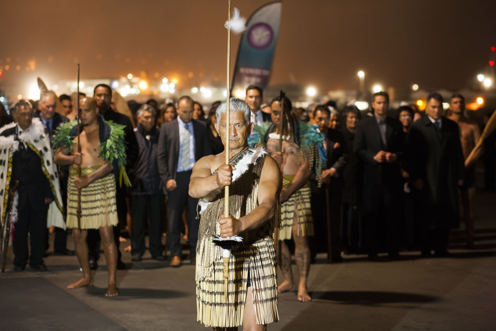 A ceremony with a Māori kaumātua in traditional dress holding a taiaha in front, two younger men behind in traditional dress with taiaha, and a crowd of people including a man in a korowhai. 