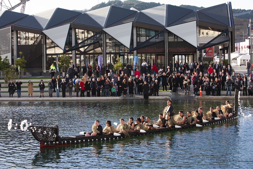 A waka with men in traditional Māori dress afloat in Whairepo Lagoon, with the Wharewaka building behind them and a crowd on the shore.