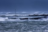 A very blue photo of severely rough seas, with waves crashing against rocks, and a small white yacht making its way through the extremely high swell.