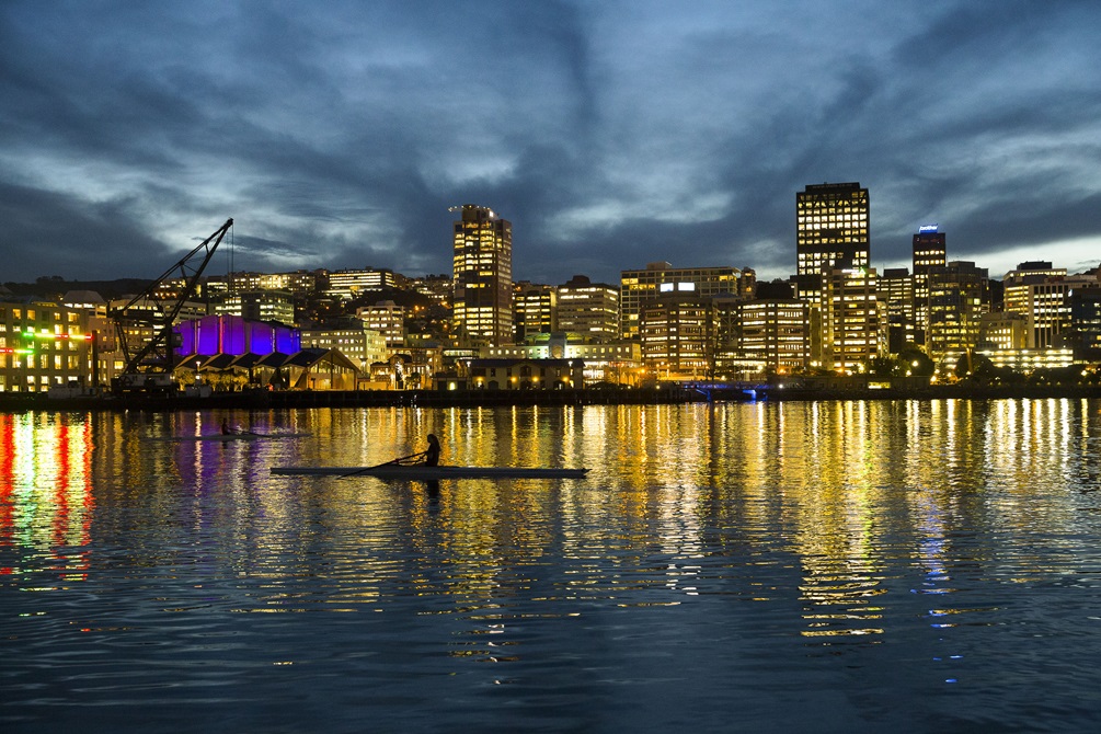 The silhouette of a lone canoe with a person in it on Wellington Harbour, with the city's buildings all lit up and reflected in the water, and a moody dark sky above.