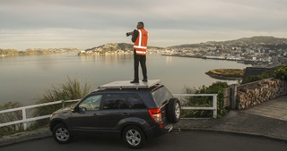 Photographer Neil Price in an orange high vis holding camera while standing on the roof of a car, looking out over Wellington harbour, with Eastbourne, Oriental Bay, and the CBD beyond.