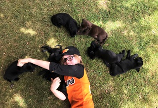 A young girl wearing a cap lying on grass surrounded by brown and black puppies.