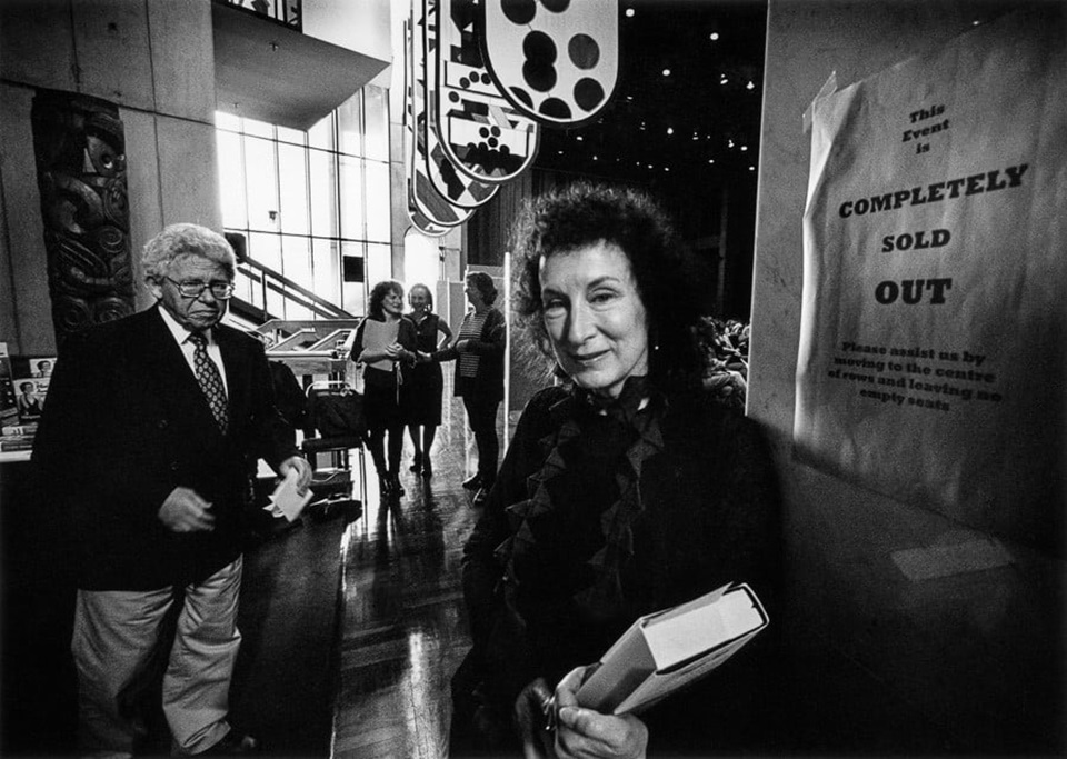 Black and white photograph of author Margaret Atwood holding a book in the Te Papa foyer with people standing in the background. 