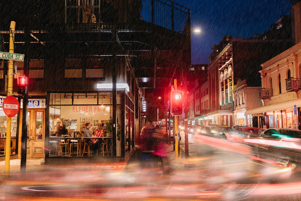 Photograph of Cuba Street at night with blurry car lights passing and a romantic couple seated at a window in a restaurant.