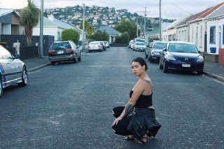 A young woman wearing hoop earings, a black tank top and black jeans, with a tattoo on her arm and back, crouching on a suburban road, with a leather jacket wrapped around her waist, and cars and houses beyond.