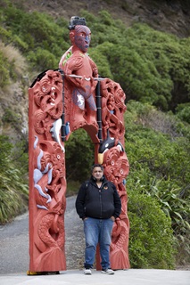 Kura Moeahu in a black hooded jersey and blue jeans, standing in front of a red, intricately carved, tall, narrow waharoa with a one metre tall tiki on top, with a gravel pathway and native bush behind.