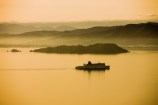 Matiu Somes Island bathed in yellow light, with mist and hills beyond, and a calm harbour with the Interislander ferry silhouetted in the foreground leaving a trail in ocean.