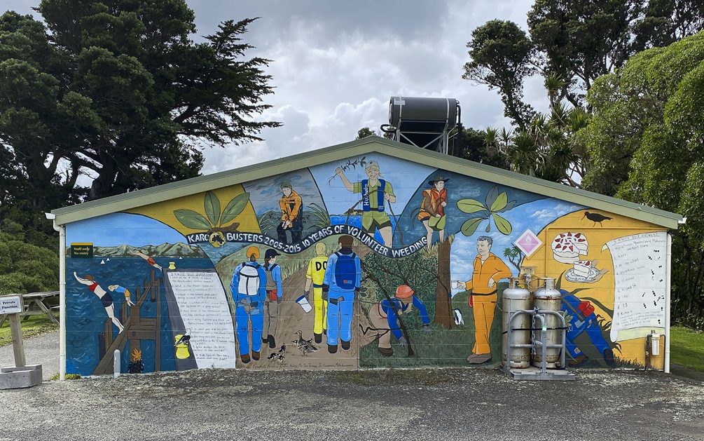 A wide colourful mural depicting people diving off a wharf and volunteers weeding in scenic environments, painted on a brick wall of a small building, with a concrete path and picnic table to the left and tall trees beyond.
