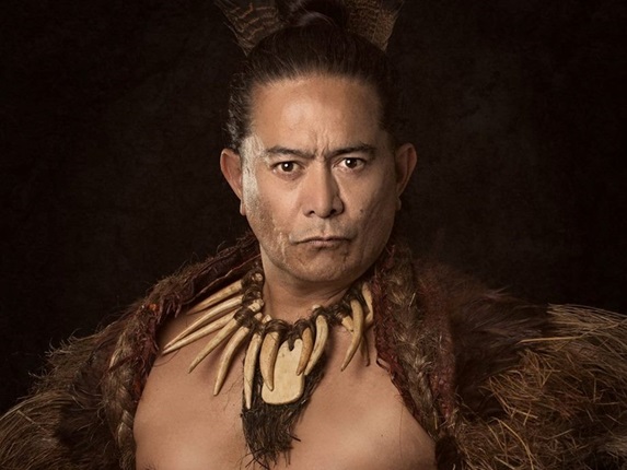 Actor and astronomer Toa Waaka, with a bare chest and looking staunch at the camera, wearing a Māori korowai and neckpiece, with a black background.