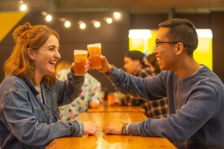 A man and a woman sitting at a long wooden table, smiling and cheersing each other with glasses of beer with the Beervana logo on them.