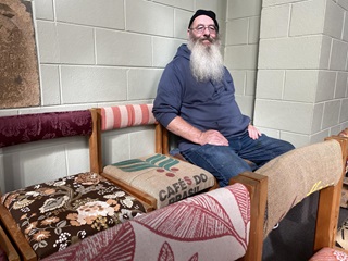 Paul with a long grey beard, a beanie, and glasses, in blue jeans and jersey, seated on a row of colourful chairs, indoors against a white cinderblock wall. 