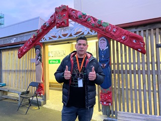 A young man doing the thumbs up in the early morning light, standing in front of a red waharoa, a Maori carved entranceway into the Wellington soup kitchen. 