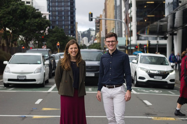 A young woman with long brown hair wearing a dark green blazer and a maroon skirt, beside a young man with shot brown hair, glasses, a blue shirt and white trousers, standing in the middle of the road with three lanes of vehicles behind them as well as tall buildings in central Wellington.