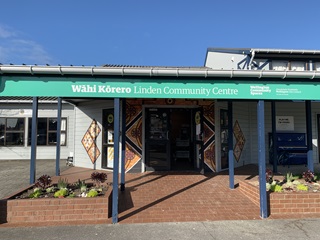 A blue sky above a wide grey building with a paved brick entranceway and garden bed on either side. There is an orange pacific-style mural around the doorway, and a turquoise and white Linden Community Centre sign under the roof. 