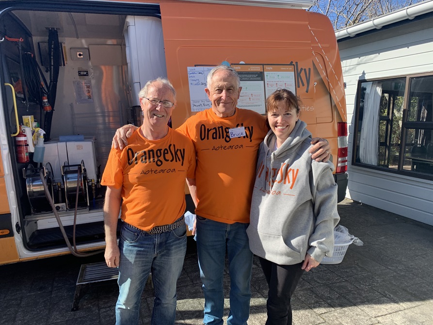 Two older gentlemen in orange t-shirts and a woman in a grey hoodie - all tops have the words Orange Sky Aotearoa on them - smiling with their arms around one another, with an orange van behind them.