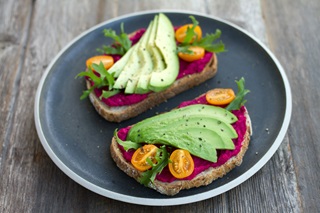 A grey plate on a wooden table with two slices of freshly-baked bread, covered in a beetroot hummus topped with perfect avocado slices, baby tomatoes, and microgreens. 