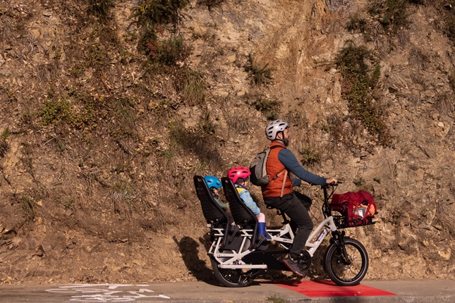 A man riding a bike that has two kids on seats secured to the back, riding uphill in a bike lane against a steep dirt hillside.