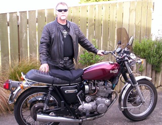 Bill Stevens dressed in black t-shirt, leather jacket and dark sunglasses, standing behind his maroon motorcycle with one hand on the handle and one hand on the seat, in front of a tall fence.