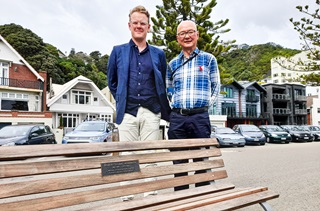 Two men smiling, standing around Oriental Bay behind a public bench seat with a dedication plaque on it, in front of a tall pine tree, houses and parked cars.