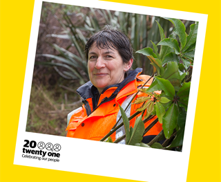 A smiling woman with short brown hair who looks to be wet, standing outside in amongst green bush wearing a bright orange high-vis waterproof jacket, framed in a polaroid with the words 20 Twenty One celebrating our people written on it.