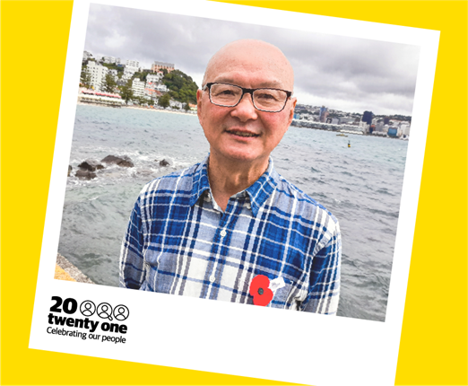 A smiling man, Bruce Tan, wearing glasses and a blue and white chequered shirt, with an ANZAC poppy pinned to it. He stands with his arms behind his back on Oriental Bay, pictured waist up, inside a yellow and white polaroid frame that says 20 Twenty One: celebrating our people.