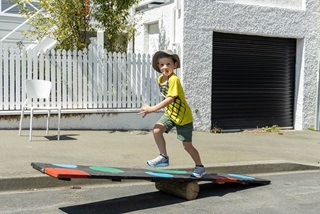 A young boy, about four-years-old, wearing a wide-brim sunhat, green shorts and a yellow t-shit, running over a colourfully-painted wooded plank that is balanced like a seesaw over a log, with a white picket fence and white garage with black roller door in background.