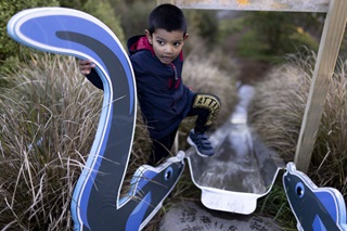 A young boy with black hair in dark blue jacket and track pants, stepping onto the top of a steep slide that has large bright blue handles at the top and is perched on a hill covered in native grass.