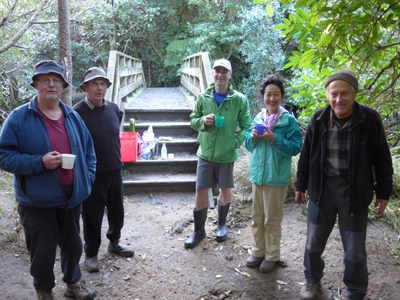 Five men and woman holding cups of tea, wearing hiking clothing, standing in a bush setting in front of steps and a wooden walking bridge.