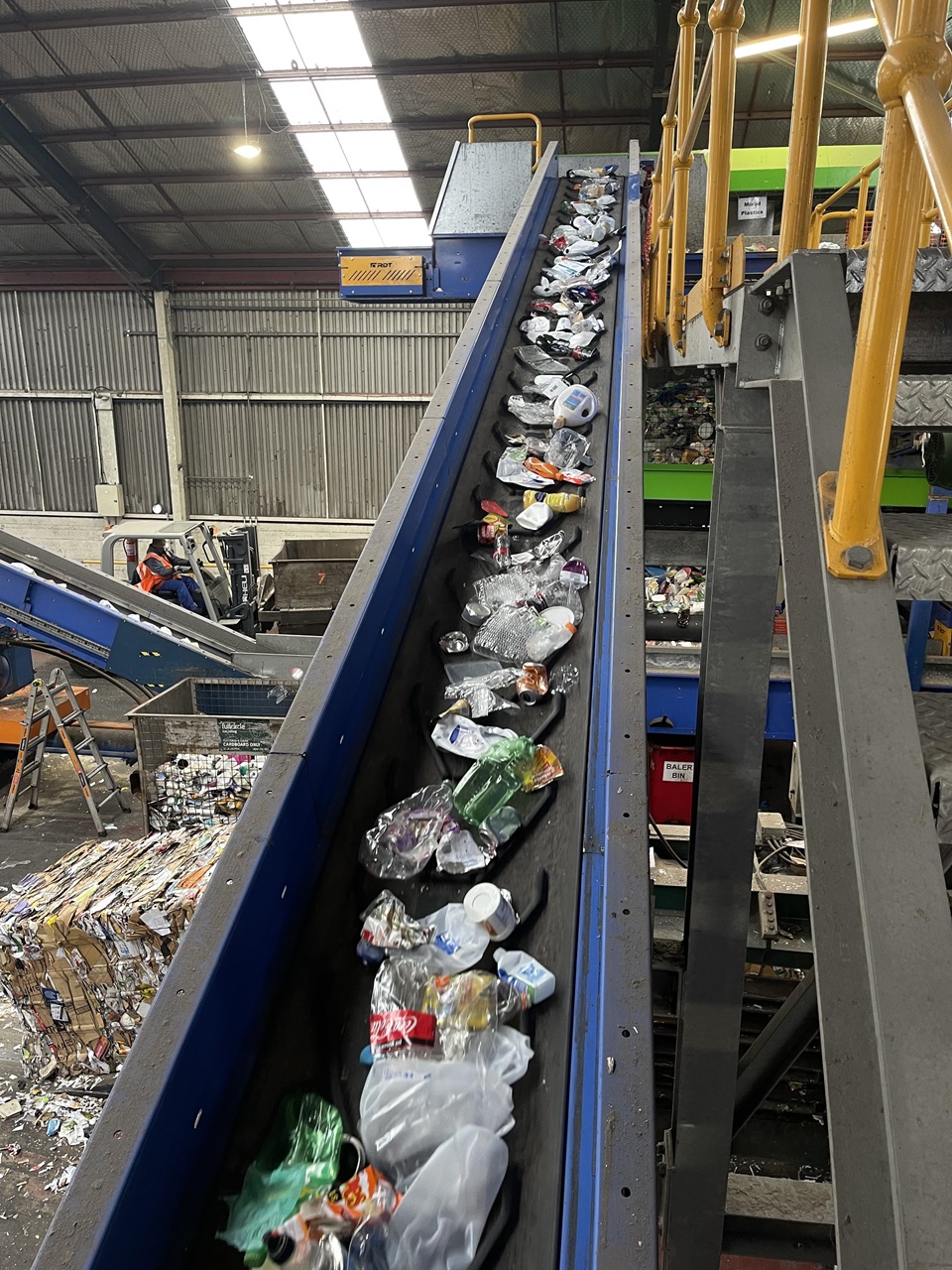 A blue conveyor belt belting plastic bottles and containers upwards, with a yellow and grey metal staircase to the right, and bales of recyclable materials being prepared below.
