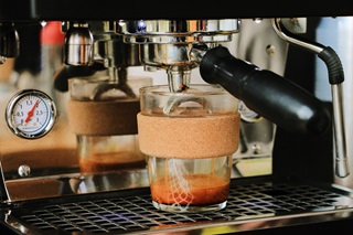 A glass and cork reusable coffee cup on a commerical coffee machine being filled with coffee. 