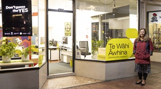 Marama Davidson standing in a skirt and red jacket to the right of the glass-windowed front of Te Wahi Awhina, a community support hub on Courtenay Place.