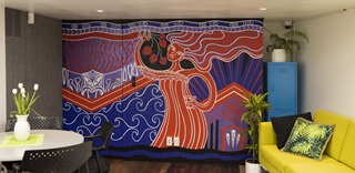 A red, blue, purple, and white mural of Māori influence on a wall at the back of a room with white flowers on a round table to the left and a yellow couch and tall blue filing cabinet to the right.