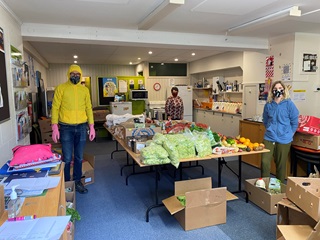 A large room with blue carpet and three people wearing masks and colourful clothes standing spaced out among tables filled with cardboard boxes and food, including fresh produce and bottles of milk.