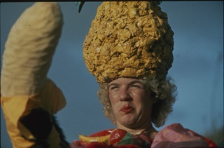 A person wearing a large yellow headdress and colourful costume makes a funny face at a giant fake banana.