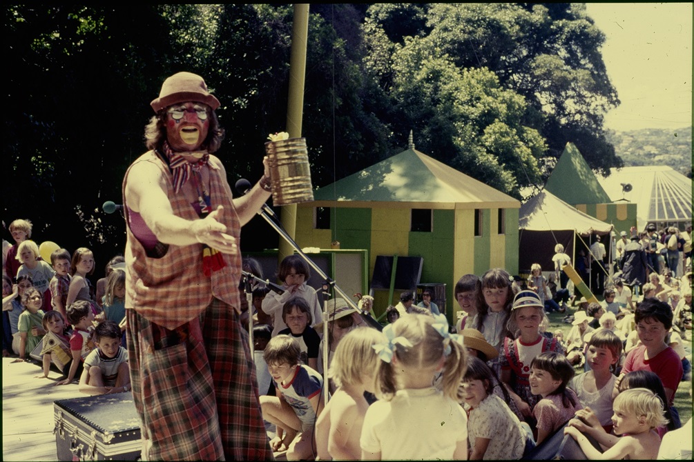 A clown holding a tin can performs for a crowd of children from a stage.