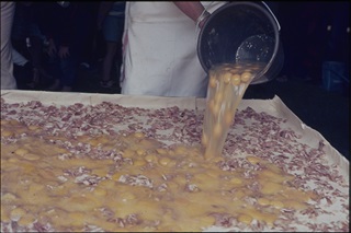 A baker pours hundreds of eggs from a bucket onto a metre long pie base covered in bacon.