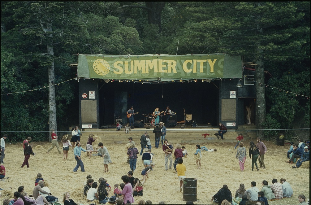 A group of people play on a field covered with hay in front of a stage with a large yellow and green Summer City banner.