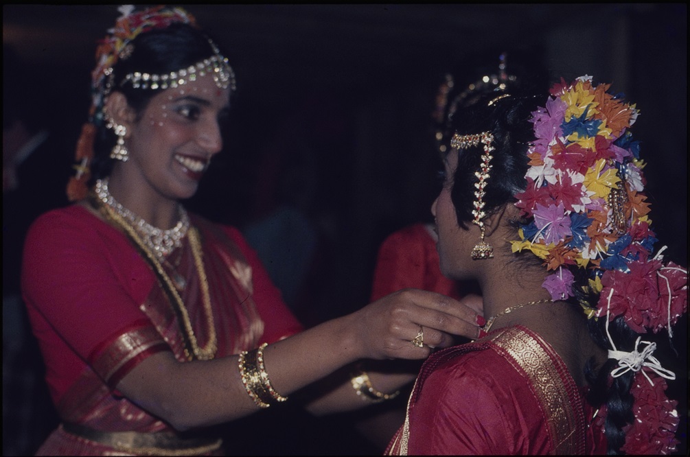 Two woman in intricate traditional Indian clothes smile at each other.