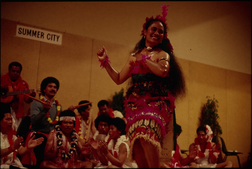 A woman dances in Pacific Island traditional dress while a group perform music behind her.
