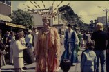 Men dressed in colourful, draping garments and large headdresses walk down a street watched by a crowd of people.