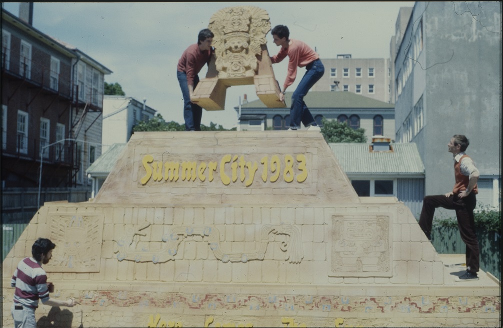 4 men work to assemble a large model of a pyramid which reads Summer City 1983 for a parade float.