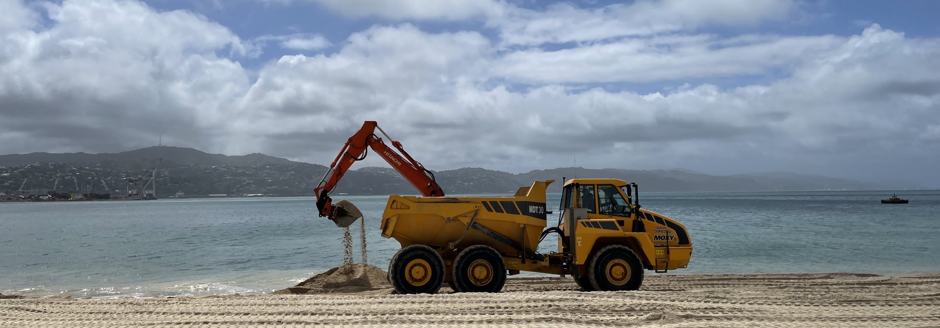 A yellow digger truck scooping up sand on the beach by the shoreline, with the water and land in distance behind and golden sand in foreground.