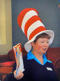Raewyn wearing a red and white striped Doctor Suess hat, reading a book from a red armchair.