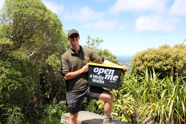 Park Ranger Adam on a sunny day, wearing his dark green uniform of shorts and t-shirt and a cap and sunglasses, posed with a leg up on a rock, holding a large black and yellow plastic container that says 'open me' in white. Surrounded with bush and blue sky above.