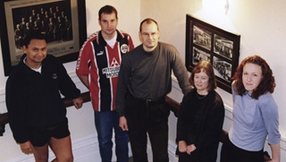 A high-angle shot of three men on left and two woman on right, standing in a row, with framed heritage photographs behind them.