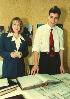 A smiling woman with a blond bob and fringe in a blue suit with white shirt and a large collar, standing beside a tall man with black hair, black dress pants, a white shirt with rolled-up sleeves and a red and black tie, standing over large planning documents and heritage books.